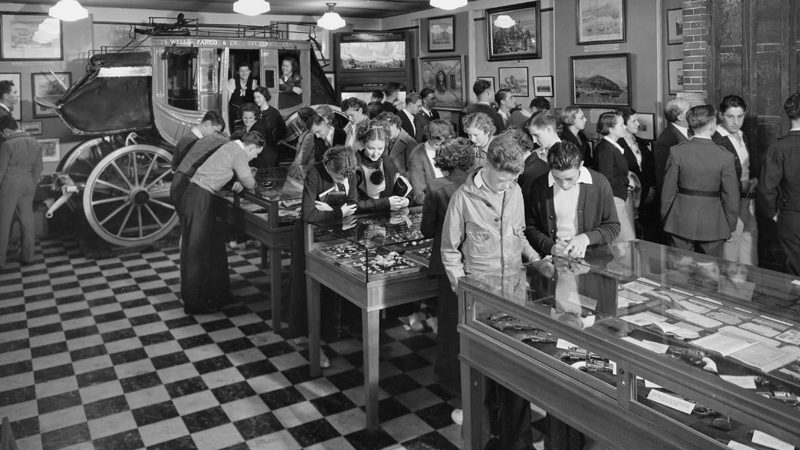 A black and white image shows a crowd of people standing to look at frames on the wall or looking down at tables with glass cases and items inside of them. At the back of the room is a stagecoach with two people inside.