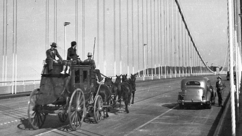 A black and white image shows a stagecoach with three people riding on top and four horses moving across a bridge. On the side of the road, a car is parked, and a man stands beside it.