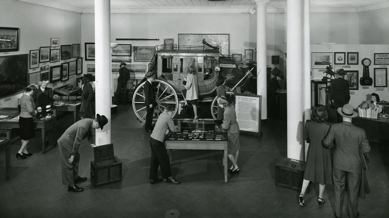 A black and white image shows a large room with column, frames on the wall, display cases, and a stagecoach in the center of the room. Around the room, 10-15 people stand to look at the displays or the stagecoach.
