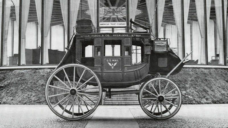 A black and white image shows the side of a stagecoach with: Wells Fargo & Co. Overland Stage written at the top and U.S. Mail on the door. It appears to be outside and in front of a window because curtains are seen.