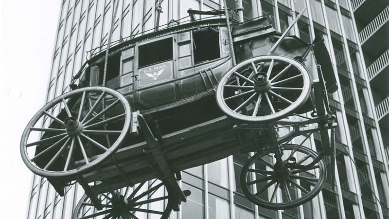 A black and white image is taken showing the bottom and side view of a stagecoach being lowered beside a building. The stagecoach has U.S. Mail written on the door.