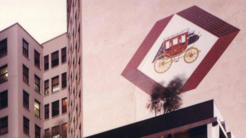 The side of a building is shown with a diamond shape in the middle. The diamond has a red border, with white and a red and gold stagecoach in the middle of it.