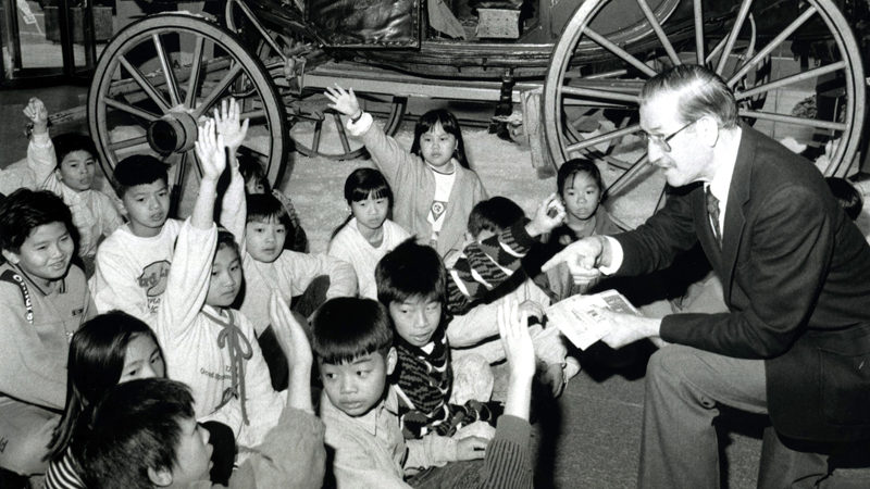 A black and white image shows part of a stagecoach and its wheels behind a group of about 15 children who are sitting on the floor. About half of them have their hands raised. A man sits in front of them, pointing and looking at one of the children.