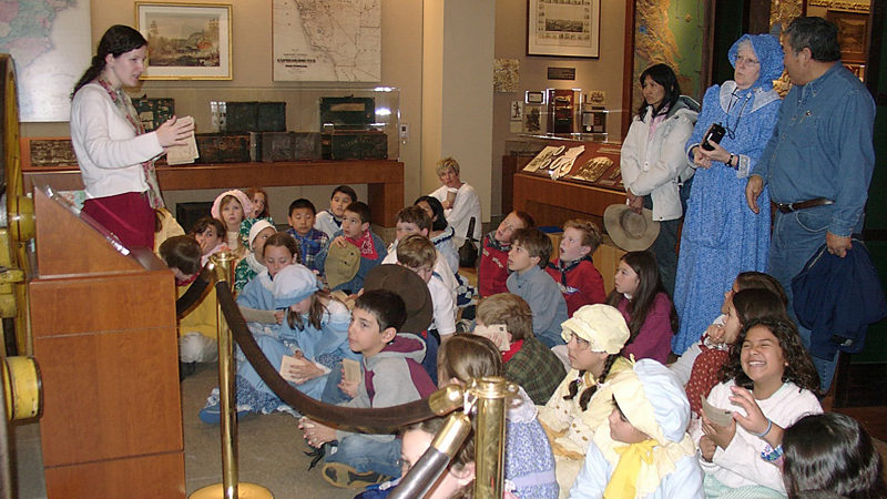 An image shows a group of about 20 kids sitting on the floor. Some are wearing bonnets. Many are looking at a woman who is standing in front of them. Several adults stand behind the children. One woman is wearing a blue dress and bonnet.
