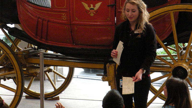 A woman stands in front of a red and gold stagecoach. Two children are seen sitting in front her, and she hands one a ticket. She has other tickets in her other hand.