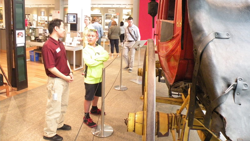 Part of a red and gold stagecoach is seen to the right of the image. To the left is a man talking to a boy as they stand nearby. Behind them, several people are looking at frames on the wall or the stagecoach.