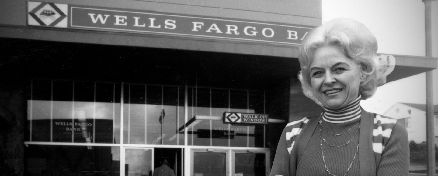 Shirley Nelson stands outside of Wells Fargo branch, smiling with arms crossed. Historic black and white photograph.