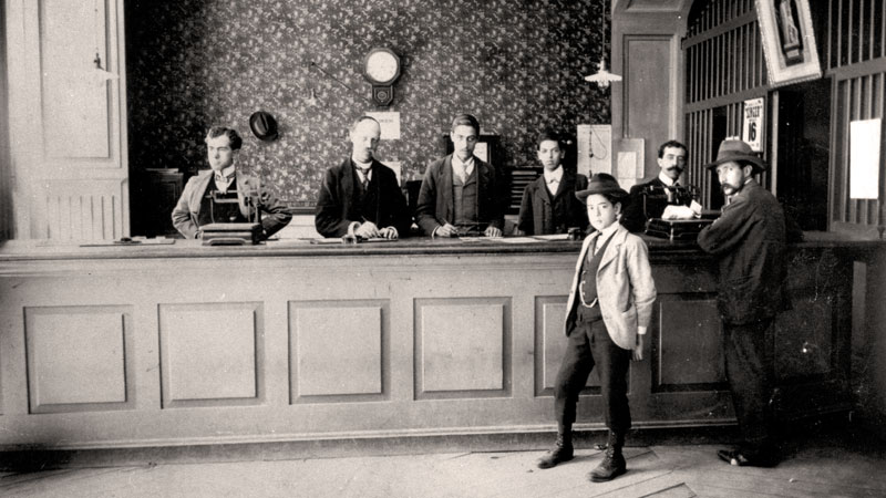 A bank teller line with five employees behind it and two people in front. Historic black and white image. Image link will enlarge image.