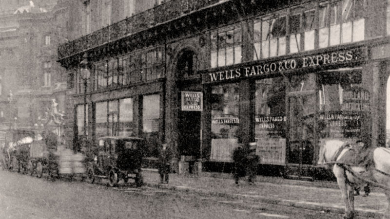 A grainy street scene showing a brick building with signage that reads Wells Fargo & Co Express. Along the street is a mixture of automobiles and horse drawn vehicles. Image link will enlarge image.