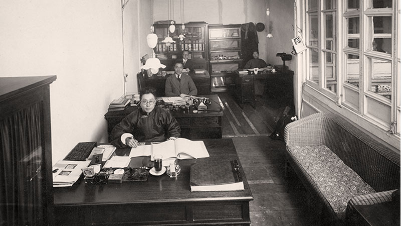 A room with three bank employees seated at desks in a row. On the right there are large windows and a small couch. The most visible desk at the front has papers, ledgers and other banking supplies across the top. Image link will enlarge image.