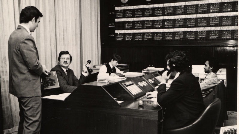 A group of employees man a telephone bank system. Behind them are large windows with white curtains and across the top of the wall hang clocks featuring the different times of international cities. Image link will enlarge image.