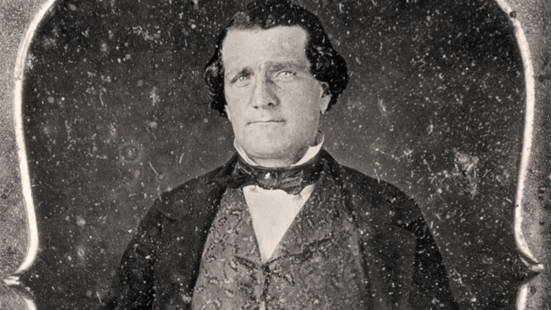 A man dressed in a black suit and bow tie with a paisley vest sits for a portrait while holding a cane. Image link will enlarge image.