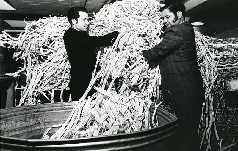 Two men in suits stand holding a huge pile of shredded paper waste above a large trash bin.