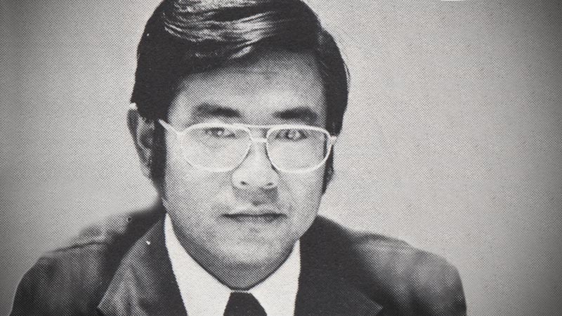 A headshot of a man with thick black hair wearing a suit and tie and glasses.