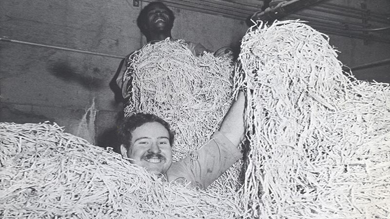 Two men’s heads peek out just above mounds of recycled paper. One man stands much higher than the other and both are smiling.