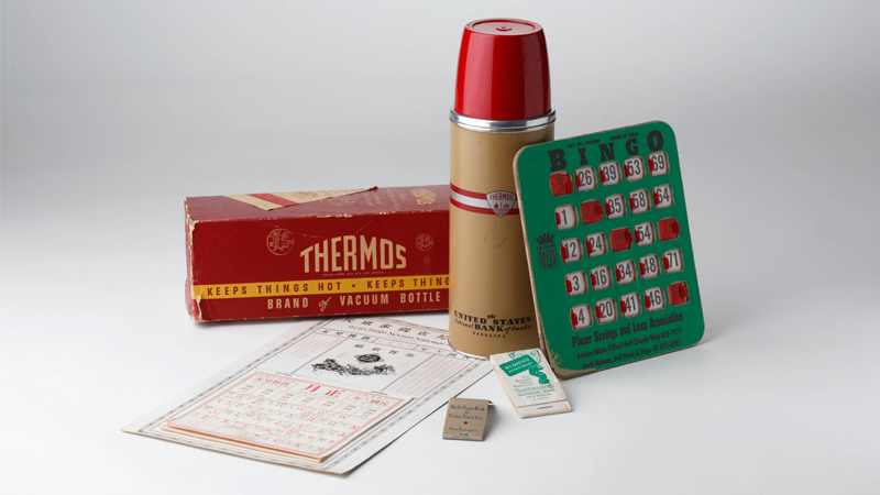 Examples of bank giveaways against a white background. It includes a red and tan thermos with original red box with yellow lettering. A green bingo card game, a deck of cards and a calendar.