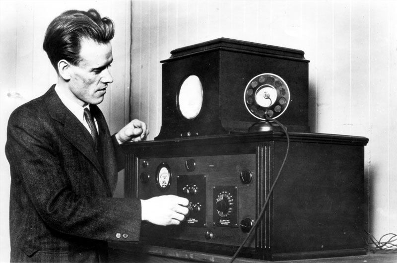 A man in a suit stands before two large black boxes with dials and microphones, which are part of the television receiver equipment set upon a small table.