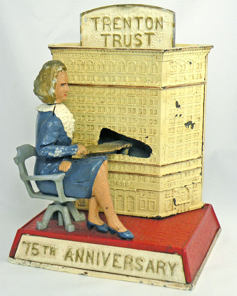 A metal coin bank with a large building in yellow and a not to scale woman seated in a banker’s chair wearing blue. Image link will enlarge image.