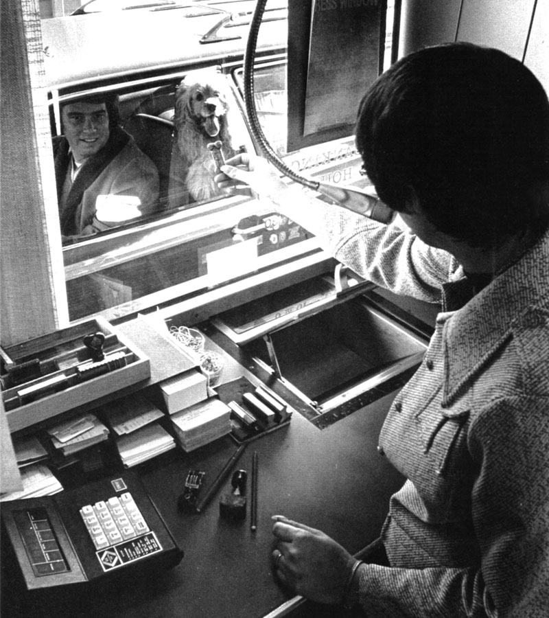 A bank teller holds up a bone shaped dog treat in view of a dog waiting for its arrival via a pneumatic tube. A man and his dog are visible in the driver’s window of the parked car. The teller’s workstation has multiple stamps, pens and adding machines. Image link will enlarge image.