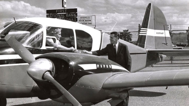 A small aircraft is parked in the foreground. From the cockpit a woman stretches out her hand to a man standing along side the plane. In the background a partial sign can be seen that reads: First National Bank of Arizona. Image link will enlarge image.