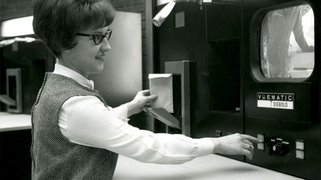 A woman teller inserts an envelope and presses buttons on a machine labeled a Vuematic. The machine allows the teller to see the drive-in customer via camera. Image link will enlarge image.