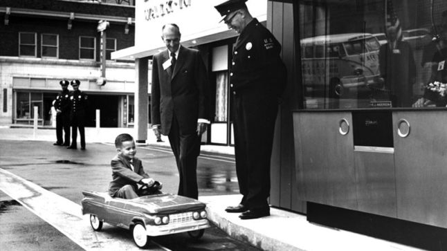 A child sits in a rideable toy car parked in a drive-in lane before a glass teller window. A bank employee and a police officer smile down on him from the sidewalk. Two more uniformed police officers stand in the background. Image link will enlarge image.