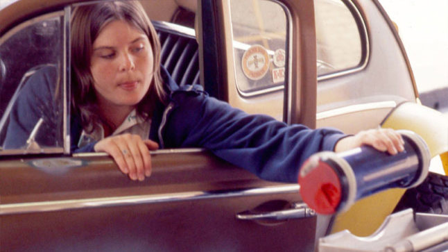 A young driver reaches out through an open door and window of their car to insert a container into a pneumatic tube system. Image link will enlarge image.