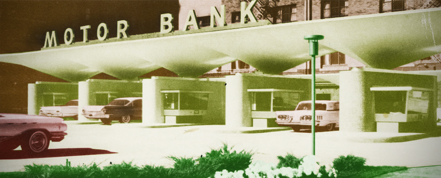 A green hued image of a 1960’s Motor bank. Three cars are parked at teller windows conducting banking business. The teller windows have glass on three sides and were occupied by employees.