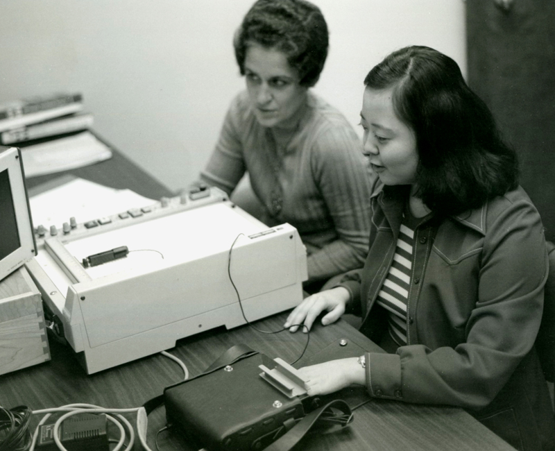 Two women seated at a table full of large computer equipment. One woman is training the other on how to use the equipment. Image link will enlarge image.
