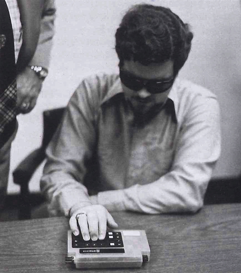A man seated at a table, wearing dark sunglasses uses a calculator designed to assist sight impaired users. Image link will enlarge image.