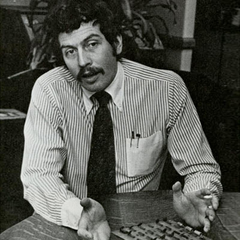 A man with dark hair and thick mustache wearing a stripped collared shirt with dark tie and pens resting in shirt pocket sits at a table with computer motherboard components.