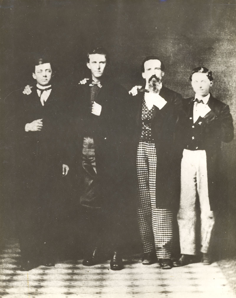Four young riders stand with their arms around one another wearing dress suits and posing for a portrait.
