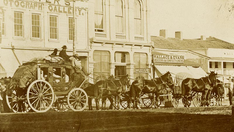 A Wells Fargo stagecoach with six horses is loaded for travel on a city street. A building in the background has a sign that reads Carters Photograph Gallery. Image link will enlarge image.