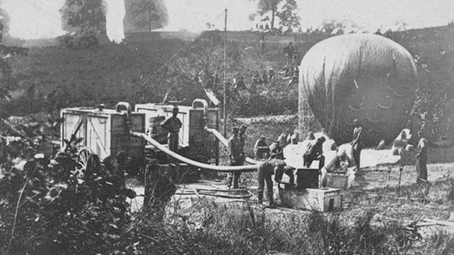 A field with men and equipment setting up and inflating a hot air balloon. Black and white historic image.