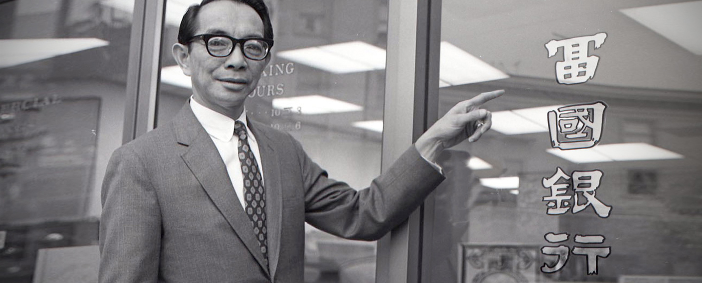 Lyman Jang in black rimmed glasses wearing grey suit, white collared shirt and printed tie. He stands outside of a bank branch. Historic black and white photograph.