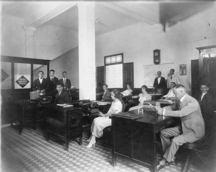 A black-and-white image shows multiple desks in a room with men and women sitting at them. Some are looking down and some are looking ahead. Image link will enlarge image.