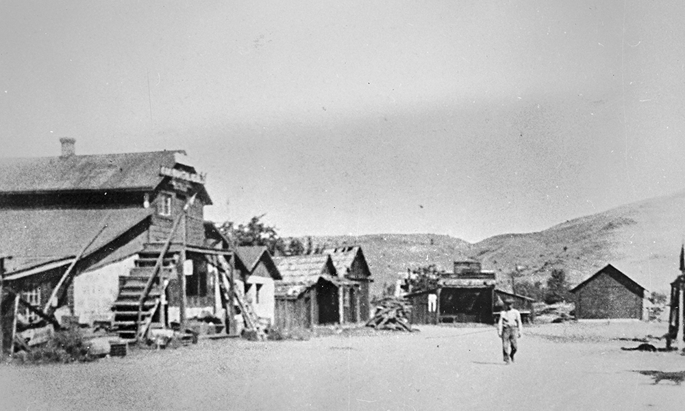 View of unpaved street with store building at left and smaller buildings in distance. Man walks in street.