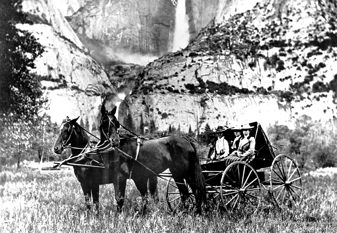 A black and white image shows a coach with two horses stopped in the middle of a field with boulders in the background. In the coach sit three women wearing dresses and hats.