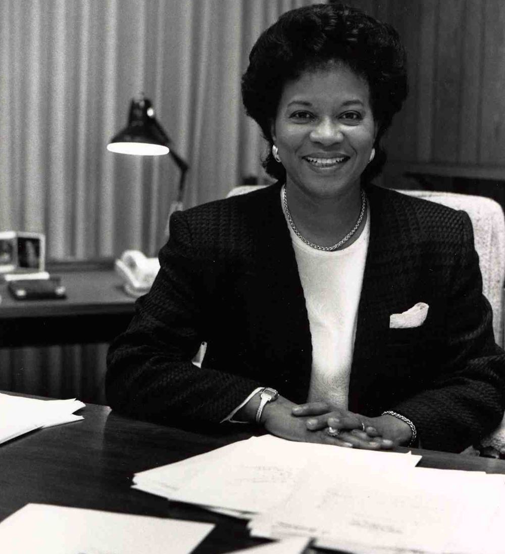 Black and white picture of a woman seated behind an office desk covered with papers.