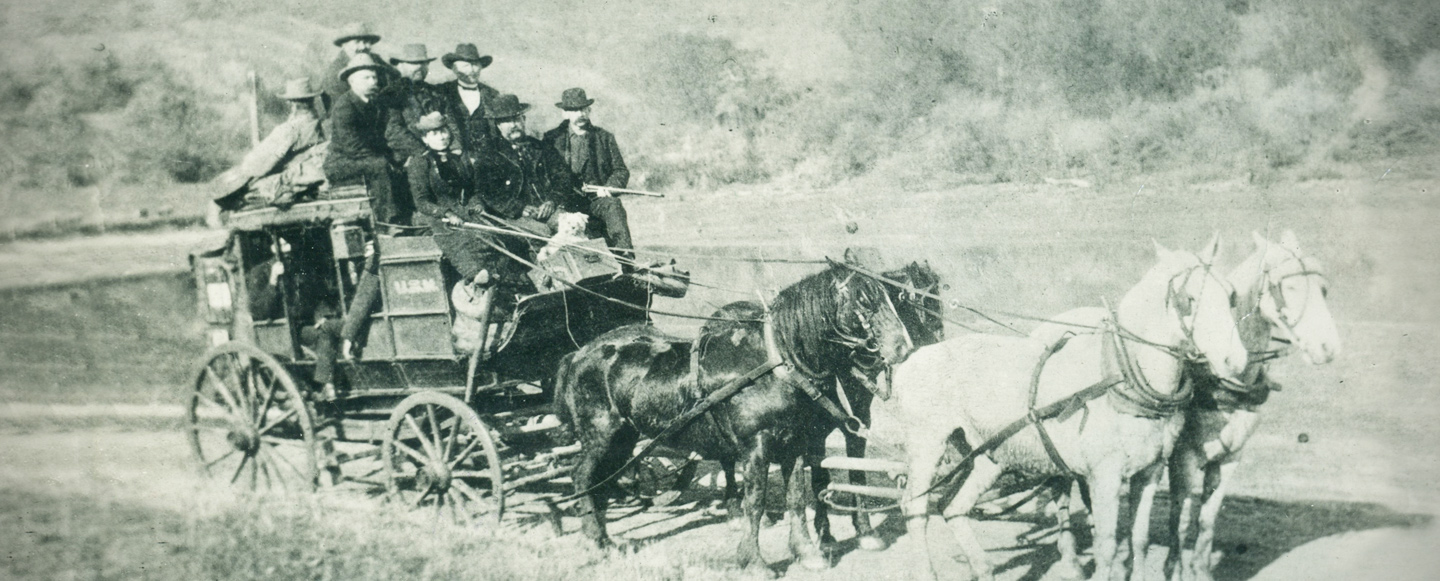 A stagecoach pulled by a team of four horses in a western landscape setting. The coach is being driven by a woman driver. There are several men seated around her on the top of the coach with additional passengers inside the coach body.