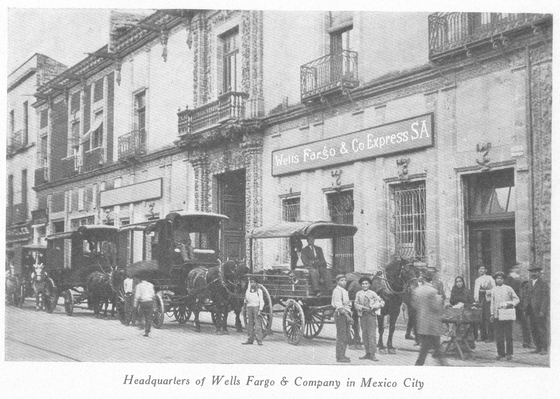 A black-and-white photo shows wagons with horses in a line outside of a row of buildings. People stand nearby on the street, and a building has a Wells Fargo & Co Express SA sign. Image link will enlarge image.