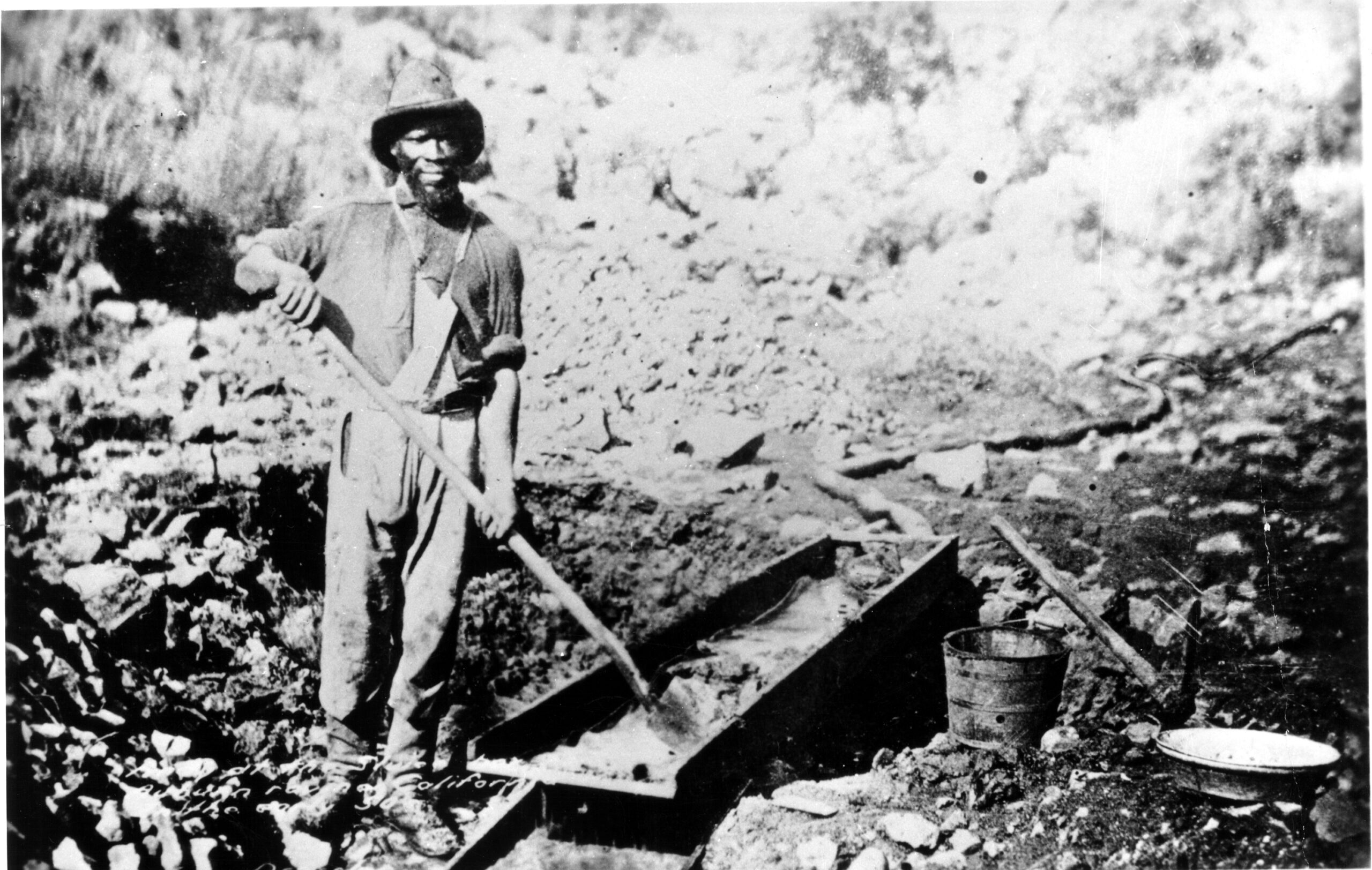 A black and white image shows a man outside holding a shovel while looking at the camera. Many large rocks are around him.