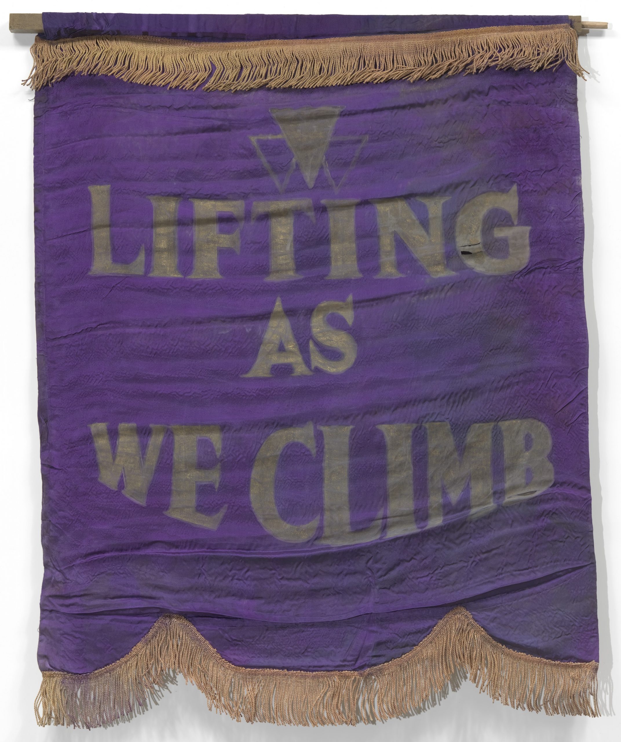 A purple cloth banner with gold fringing at the top and bottom that reads: Lifting As We Climb in gold lettering.