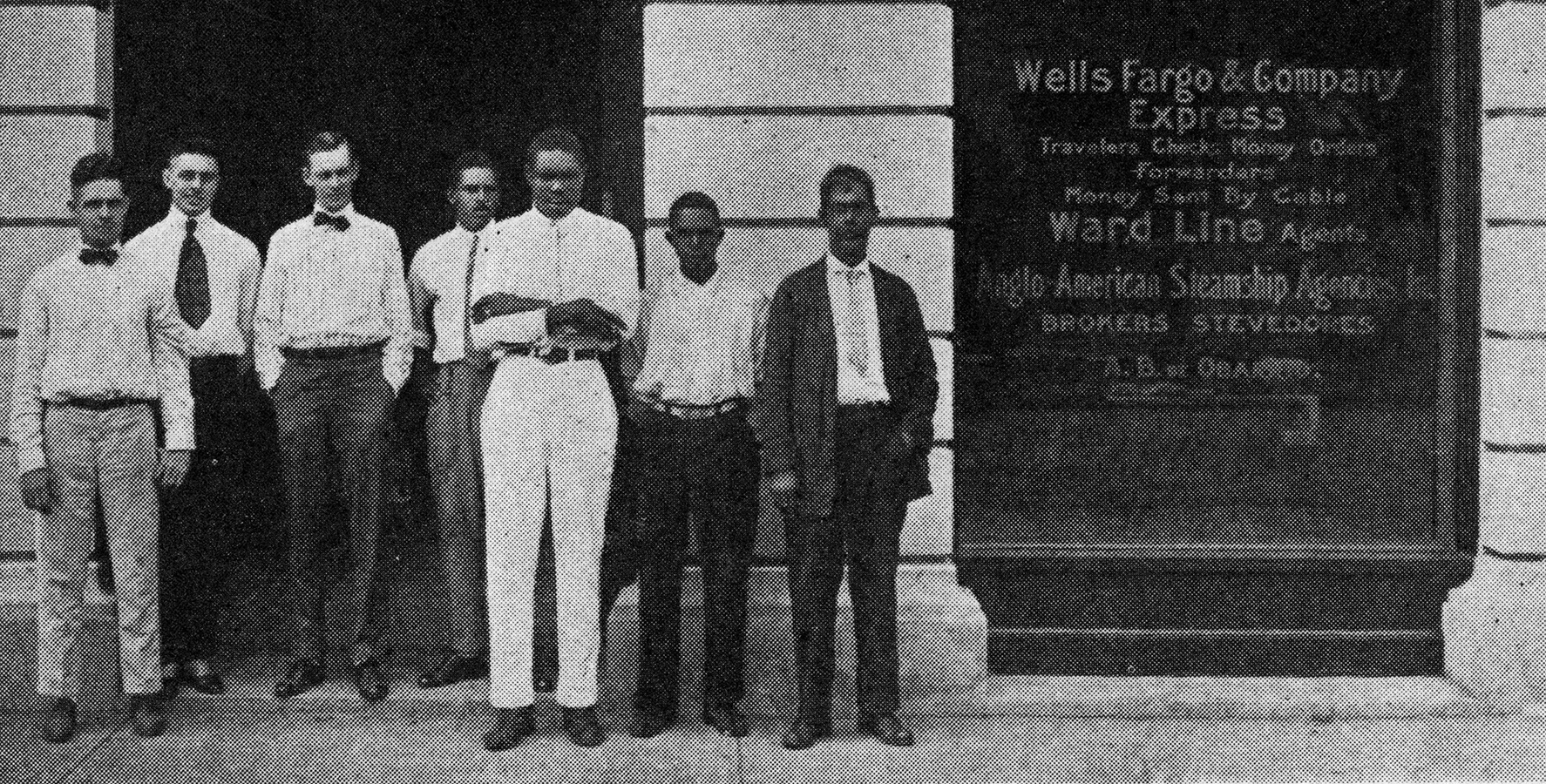 A black-and-white photo of seven men standing outside a building with Wells Fargo & Company Express on the window beside them. Image link will enlarge image.
