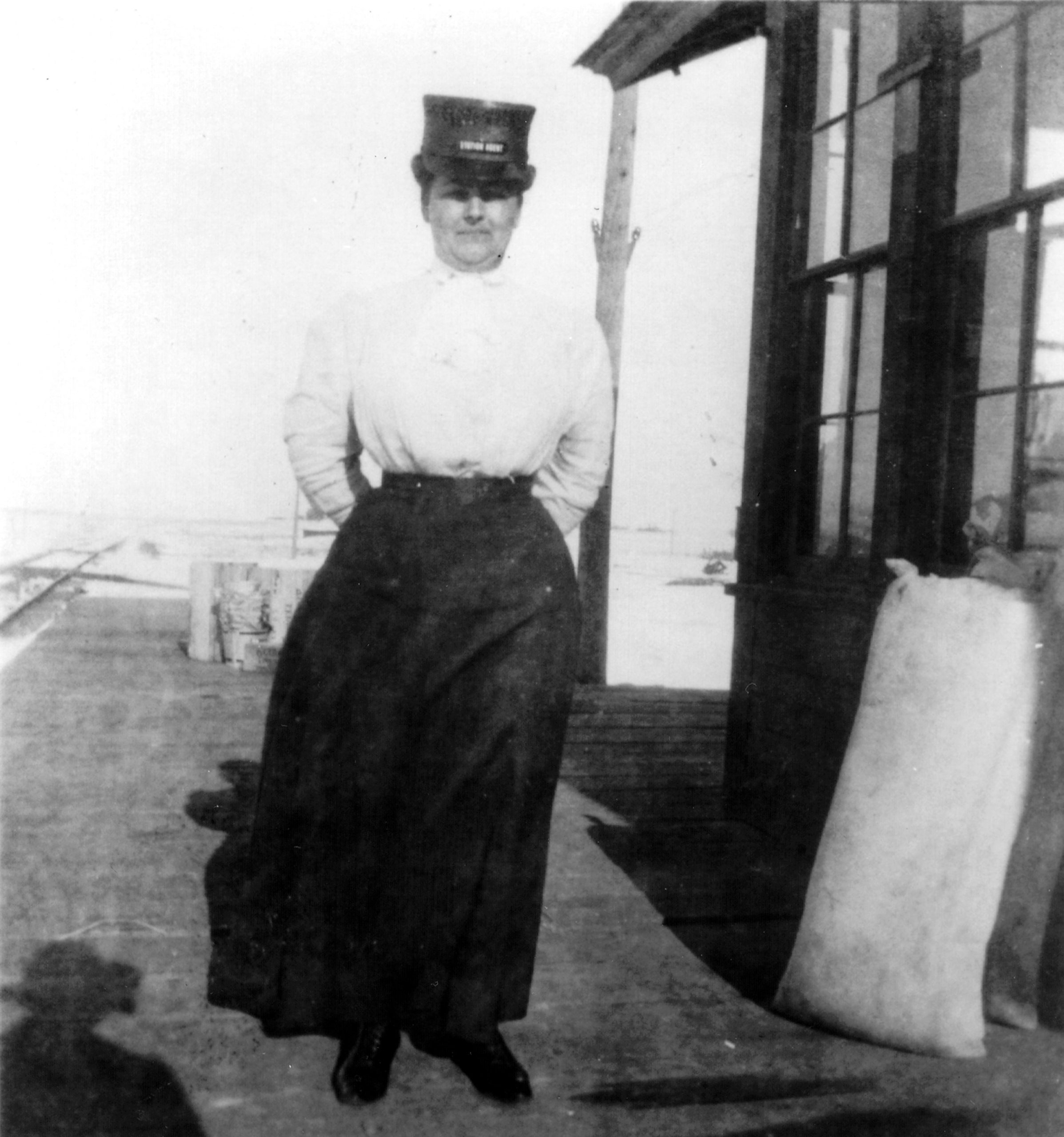 A black and white photo shows a woman wearing a blouse, skirt, and hat as she stands between train tracks and a building. Her hands are in her pockets, and full sacks are propped up against the building. A shadow is near her feet.