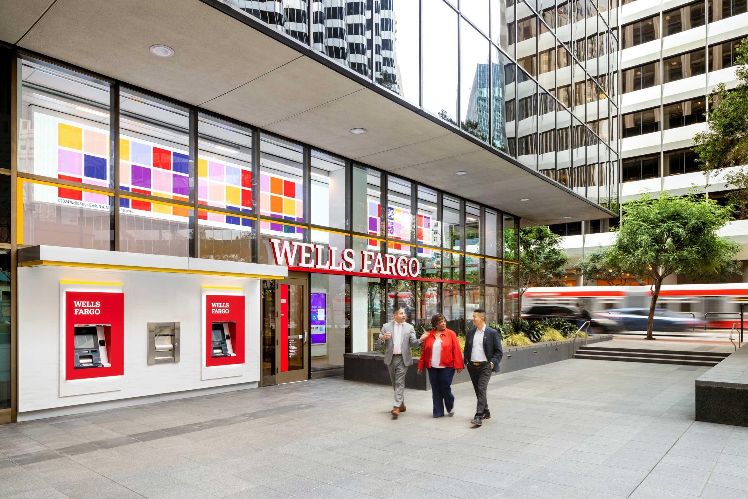 Wells Fargo branch exterior on busy street corner. Three people talking together walk past the entrance.