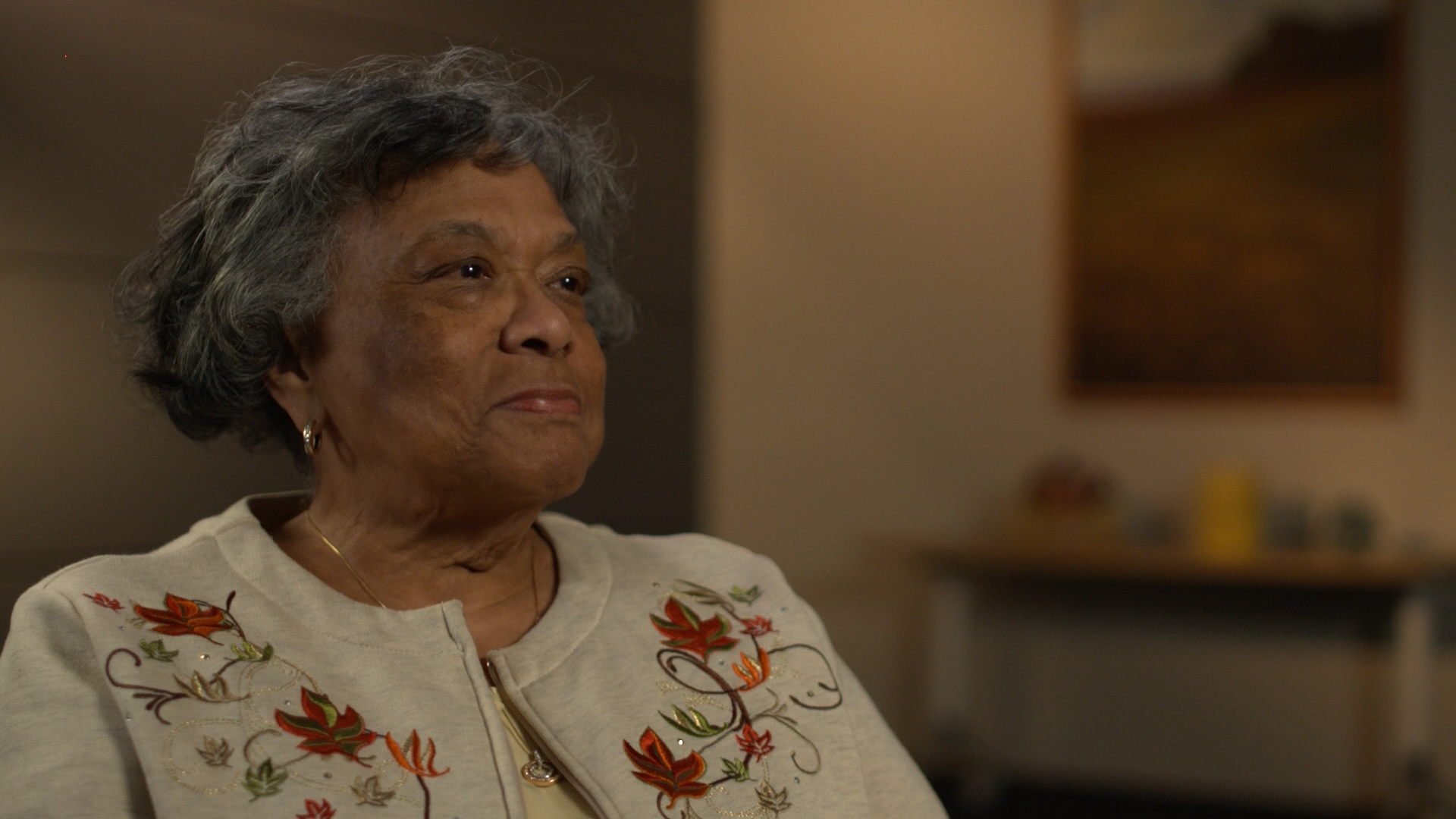 Woman seated in office smiles while looking into the distance. She has gray hair and wears a sweater with embroidered leaves.