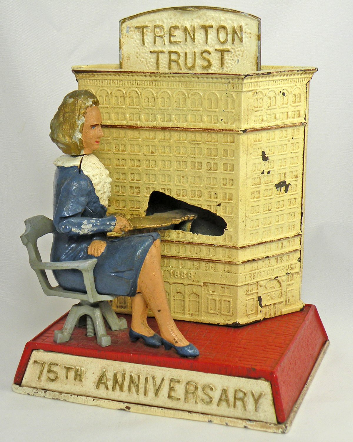 Color picture of a coin bank featuring a woman in a blue suit seated next to a building with a Trenton Trust sign. At the bottom is a sign saying the bank was made for the bank’s 75th anniversary.