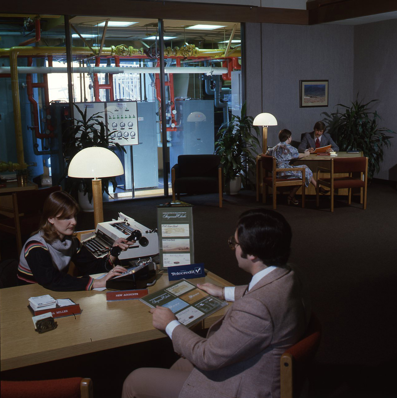 People seated at desks. In the background, red tubing is seen behind a glass wall.