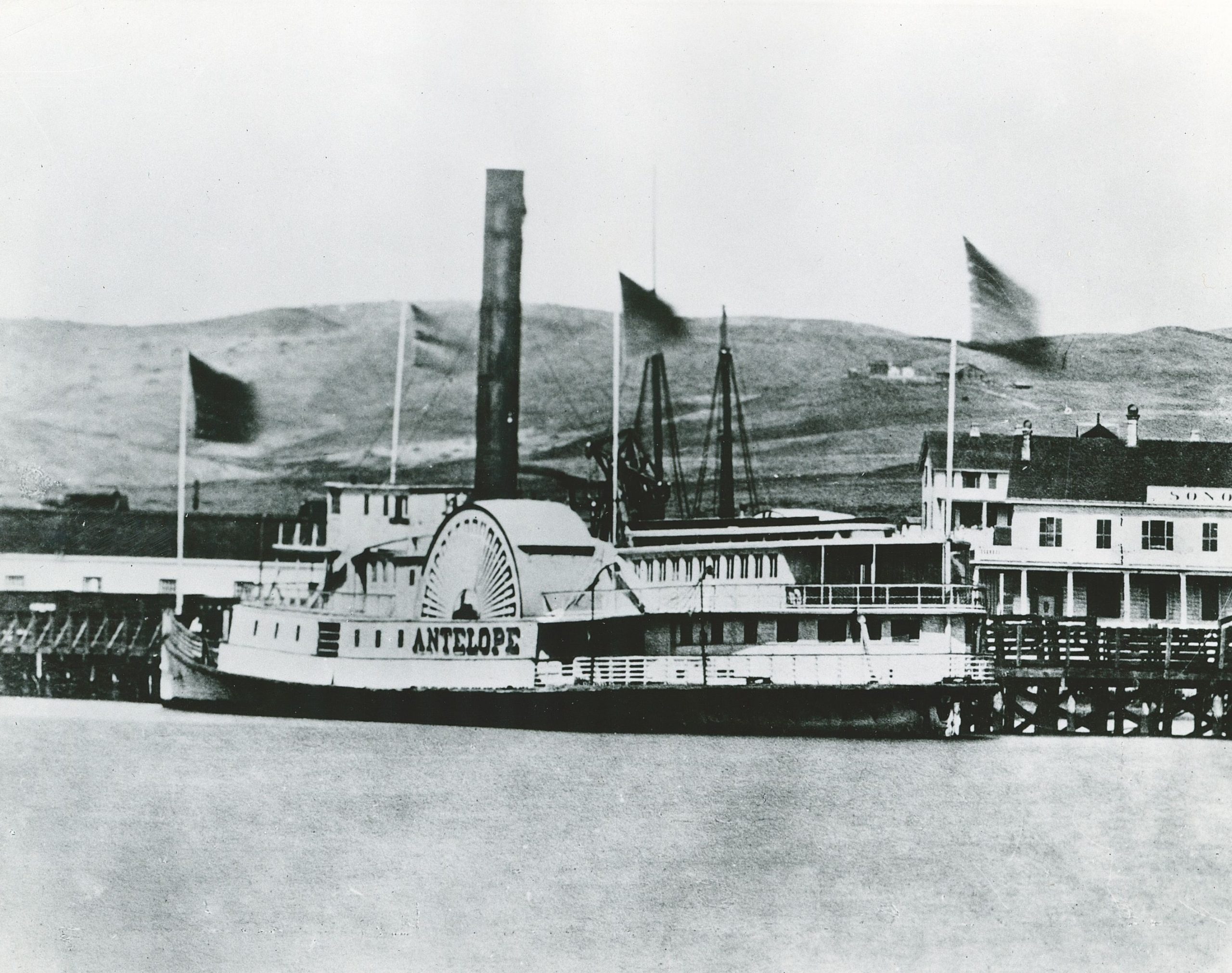 A black and white photo shows a large ship docked at a wharf with the bow pointed to left. The name Antelope is prominent on its paddlewheel on the port side. Hills are in the background, and the wharf building is to the right.